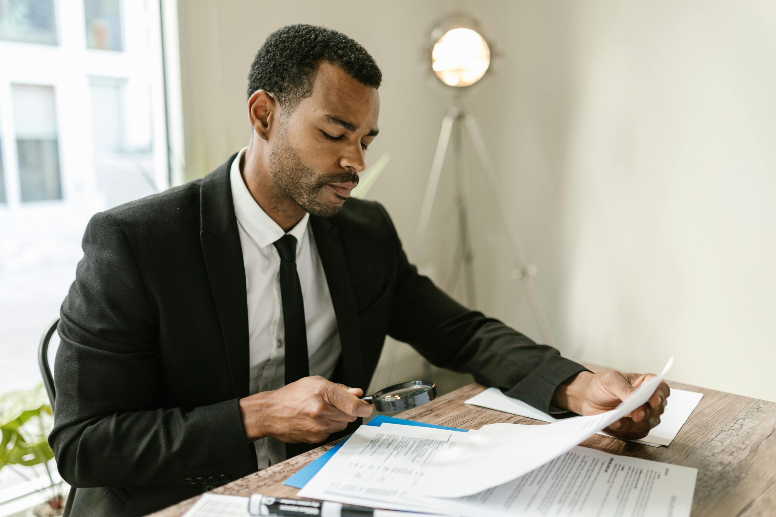Professional man examines paperwork with magnifying glass at office desk, highlighting attention to detail.